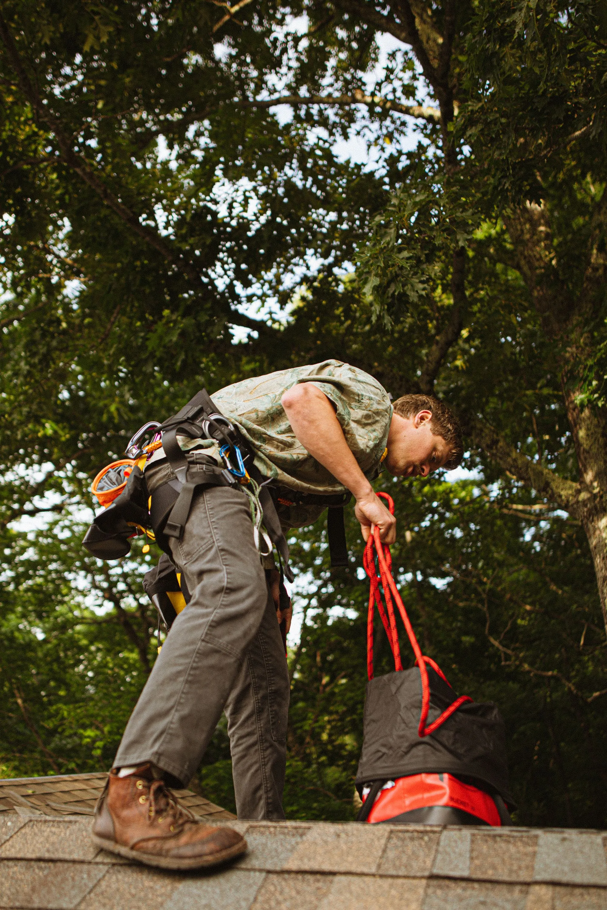 A roofer in a safety harness handling a rope line on a roof among tall trees