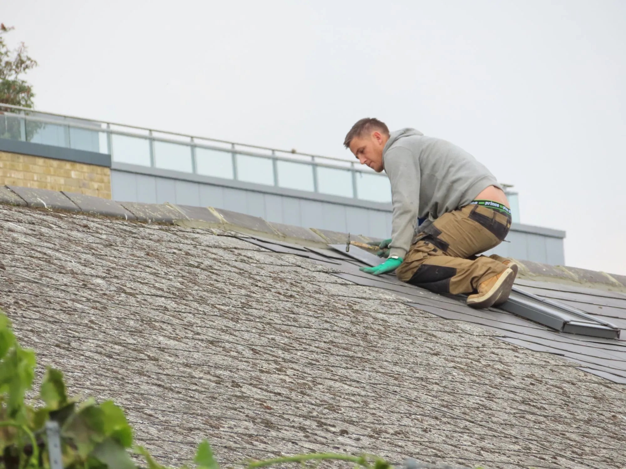 A roofer kneeling on a grey slope beside a low-pitched section of roof