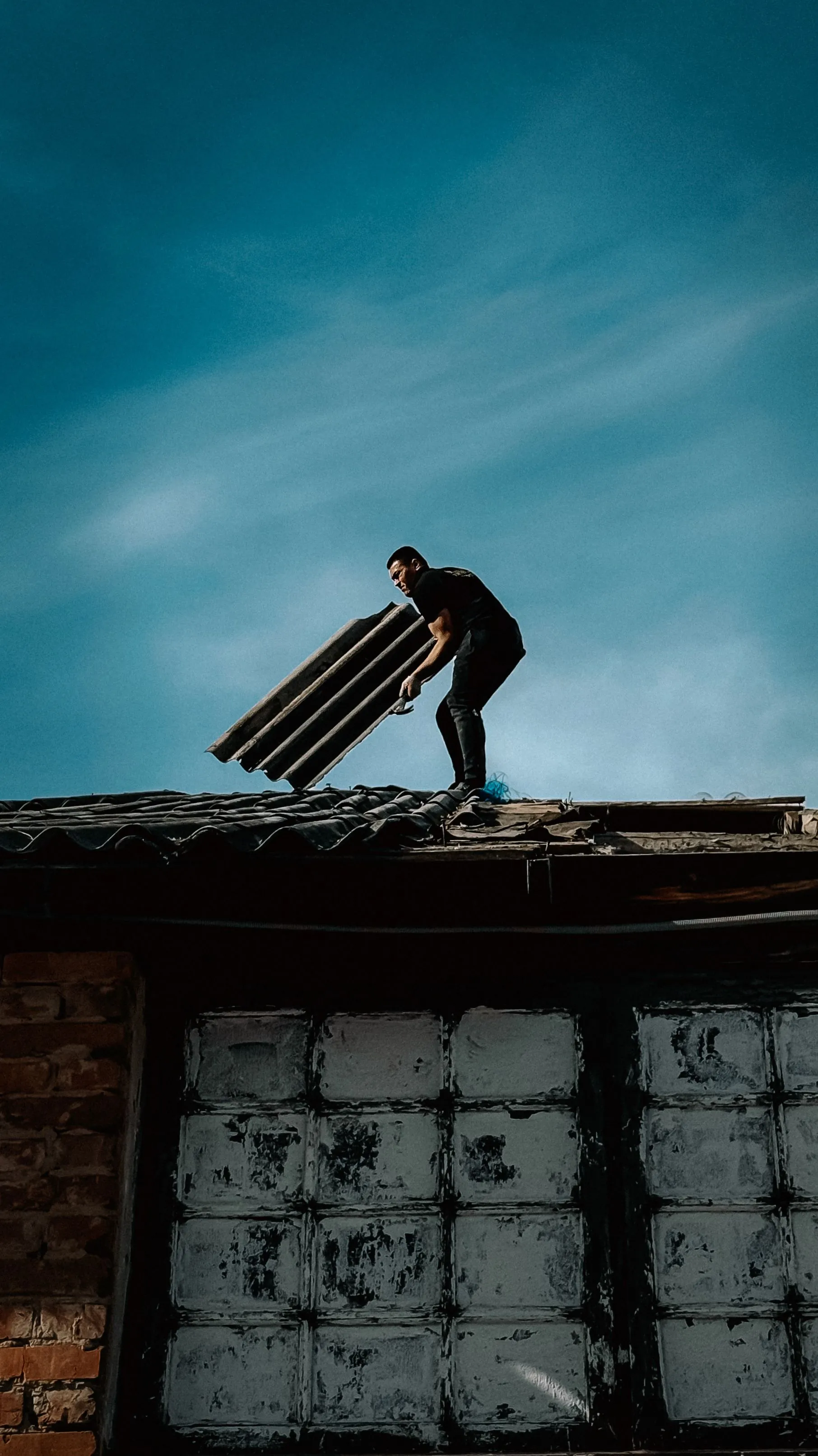 A roofer carrying a bundle of shingles across a stripped roof deck at dusk