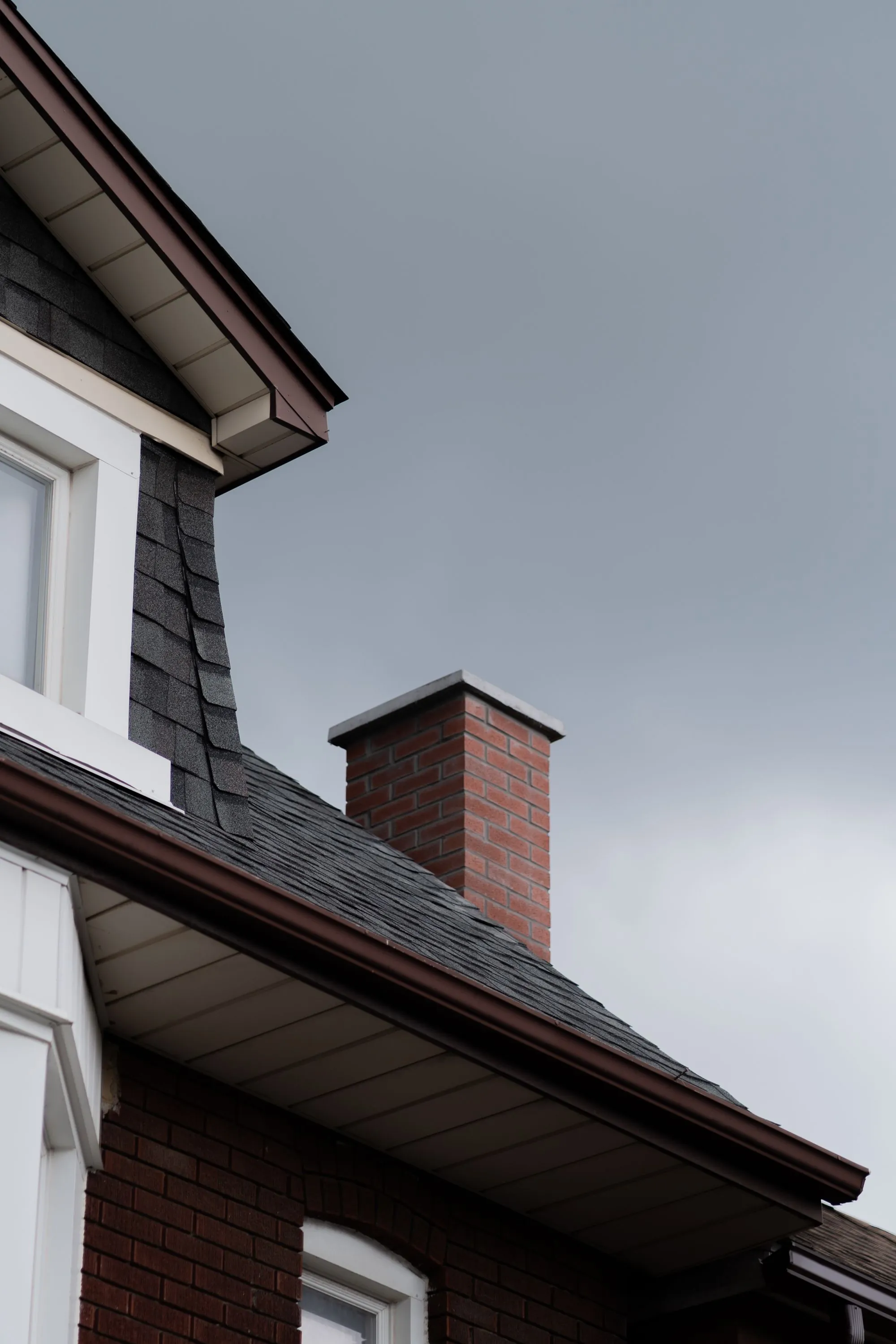 An asphalt shingle roof and brick chimney on a brick house under grey sky
